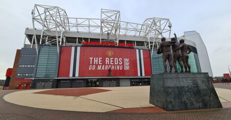 A wide view of Old Trafford, home of Manchester United, showing the stadium’s main exterior with large red banners and signage. In the foreground stands the Trinity statue of George Best, Denis Law and Bobby Charlton, set against the open forecourt and overcast sky.