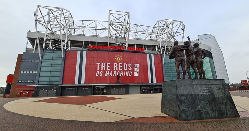 A wide view of Old Trafford, home of Manchester United, showing the stadium’s main exterior with large red banners and signage. In the foreground stands the Trinity statue of George Best, Denis Law and Bobby Charlton, set against the open forecourt and overcast sky.
