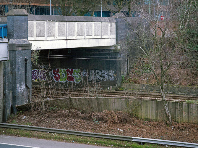 View under the bridge showing the railway line passing through a concrete underpass with brick supports.