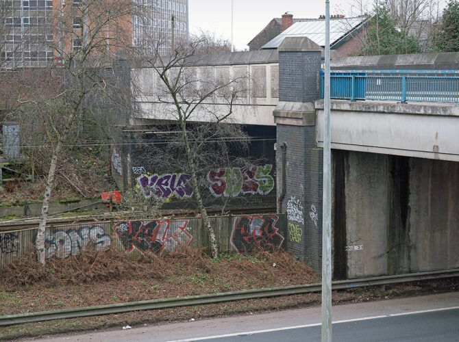The bridge spans the cutting where trains run below street level, with urban layers of road, rail and retaining walls.