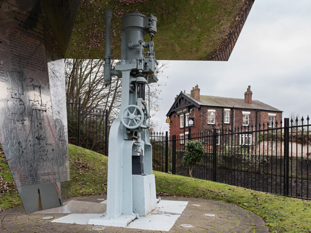A preserved steam hammer displayed beneath a metal canopy, marking the historic site of the Bridgewater Foundry. Queens Arms pub in the background