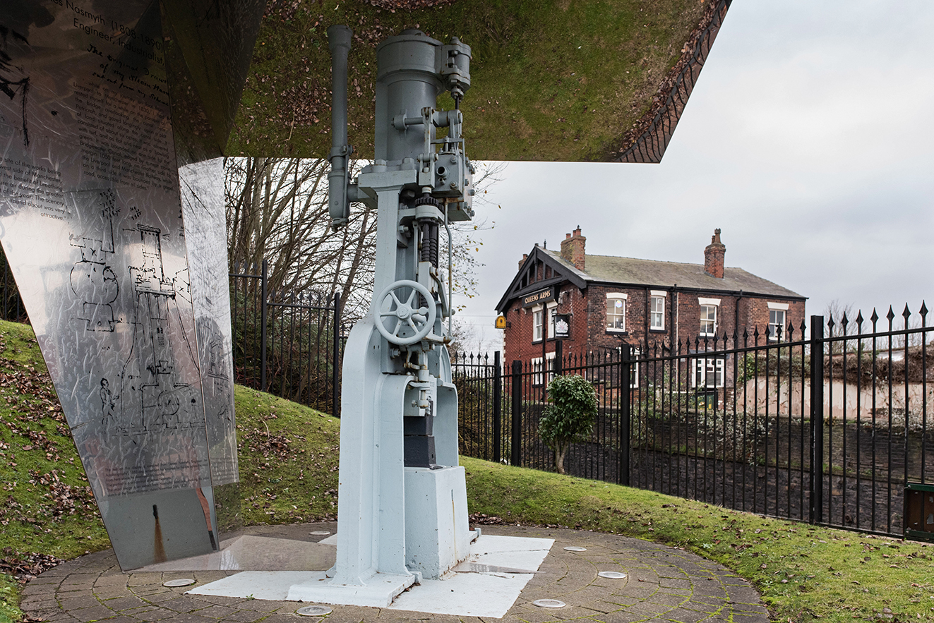 A preserved steam hammer displayed beneath a metal canopy, marking the historic site of the Bridgewater Foundry. Queens Arms pub in the background