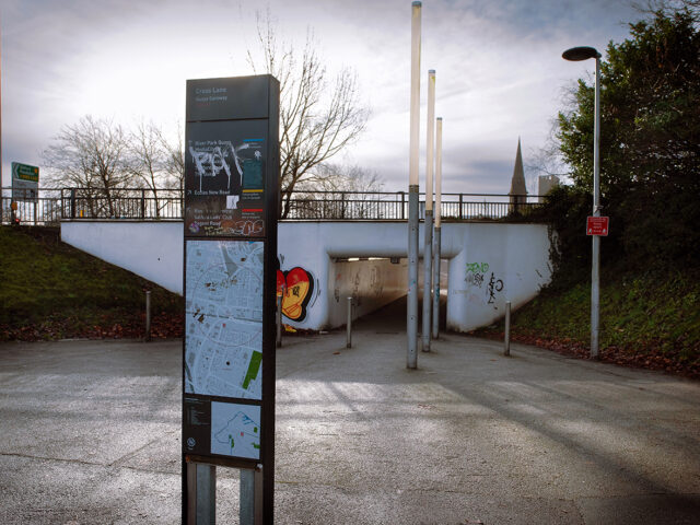 Information sign standing in front of the underpass entrance, showing local directions and map, with the road and rail bridge above