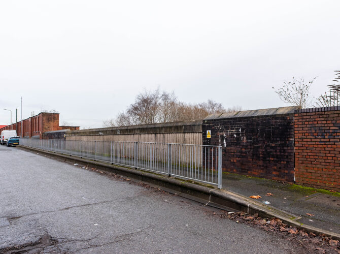 Wide roadside view showing a continuous wall with a darker brick section and a mounted warning sign, with industrial buildings and parked vehicles nearby