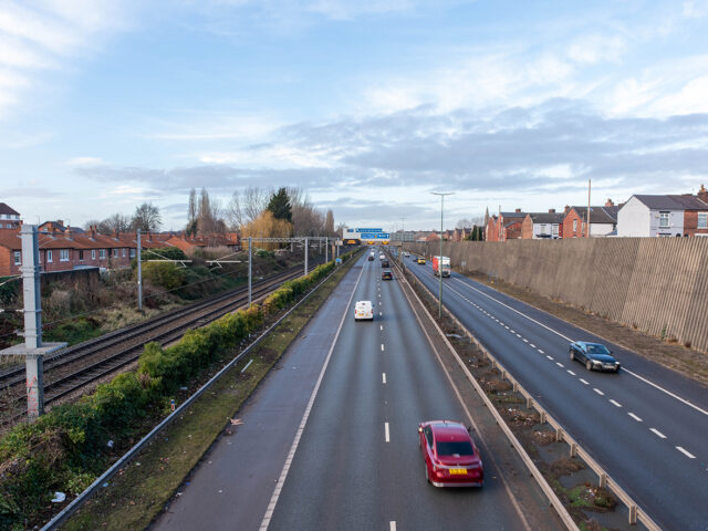 View from Gore Booth Bridge showing a railway line running parallel to a busy dual carriageway, with houses on one side and a high retaining wall on the other, marking the area where Seedley Station once stood