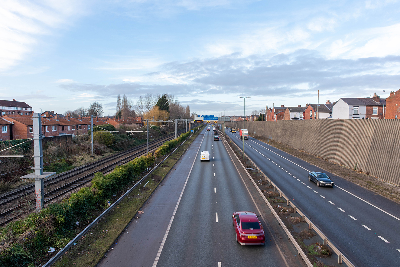 View from Gore Booth Bridge showing a railway line running parallel to a busy dual carriageway, with houses on one side and a high retaining wall on the other, marking the area where Seedley Station once stood
