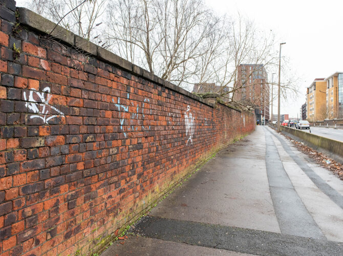 Long red brick railway wall runs beside a narrow pavement with flats and trees ahead