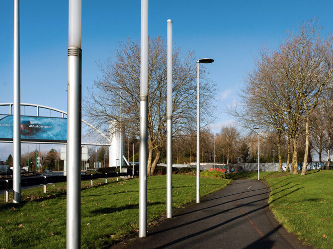 Narrow pedestrian path running through grass and trees, lined with tall metal posts and adjacent to the busy road and roundabout