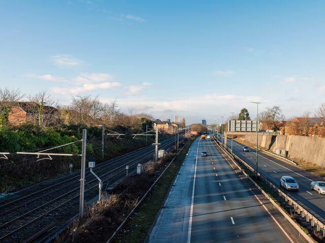 View from a bridge showing railway tracks in a cutting running parallel to a busy dual carriageway, with houses and greenery on one side