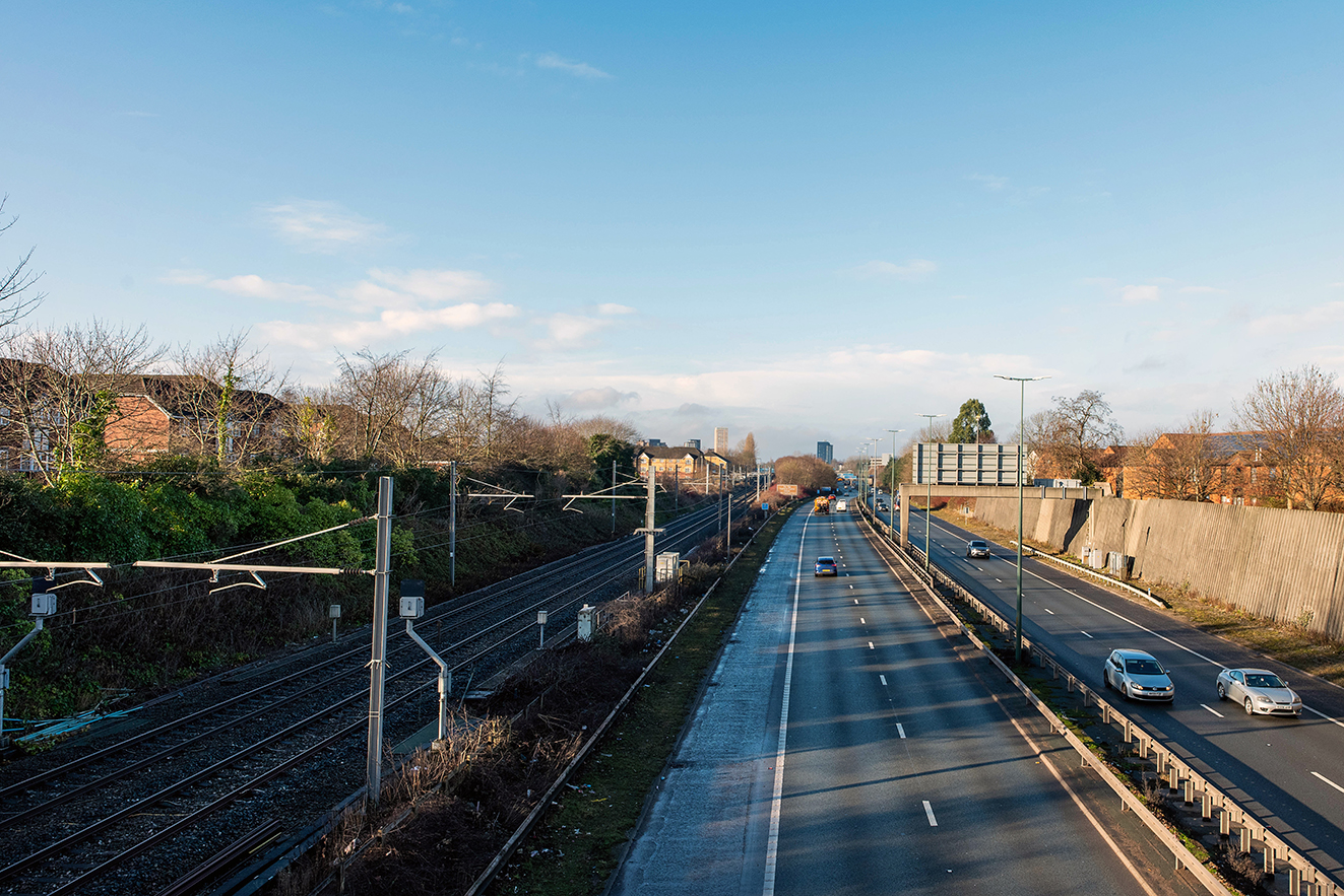 View from a bridge showing railway tracks in a cutting running parallel to a busy dual carriageway, with houses and greenery on one side