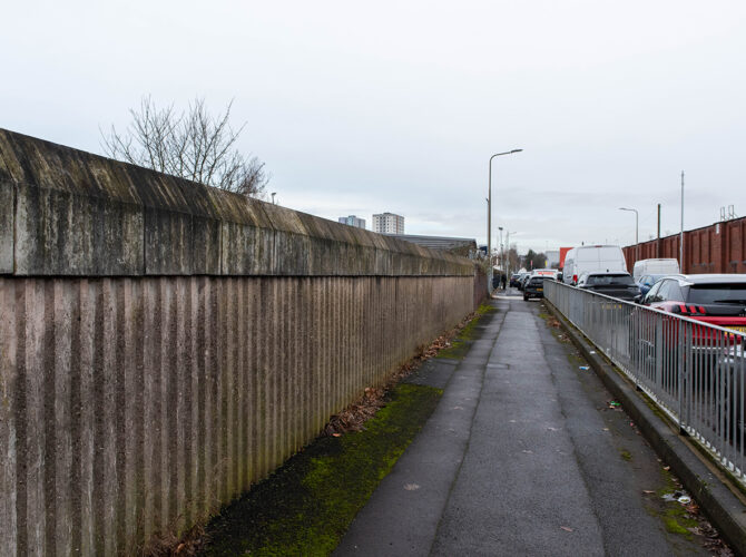 Long view along a pavement bordered by a weathered concrete wall, with moss at the base and cars parked along the road