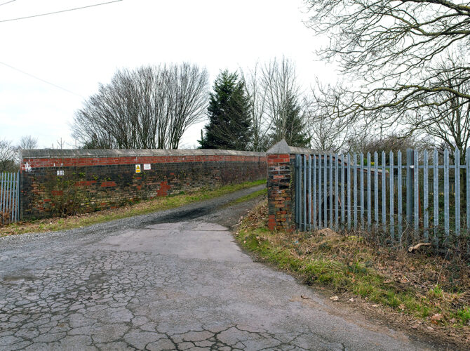 The approach to Newton’s Bridge from a paved lane, showing brick parapet walls curving inward and metal fencing to the side. The rural setting and gentle slope highlight its integration into the landscape.