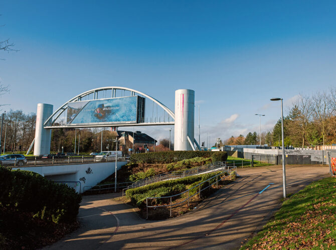 Sloped pedestrian path with railings leading down towards the underpass, with the road bridge and signage structure visible above