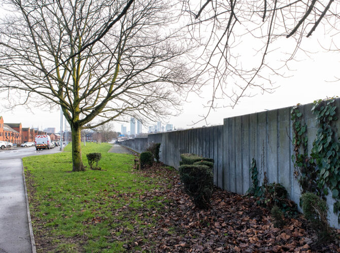 View along a grassy verge and footpath with a continuous concrete wall marking the railway cutting