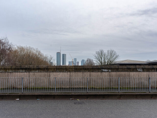 Straight-on view of a tall concrete wall behind a roadside barrier, with distant high-rise buildings visible above the top edge