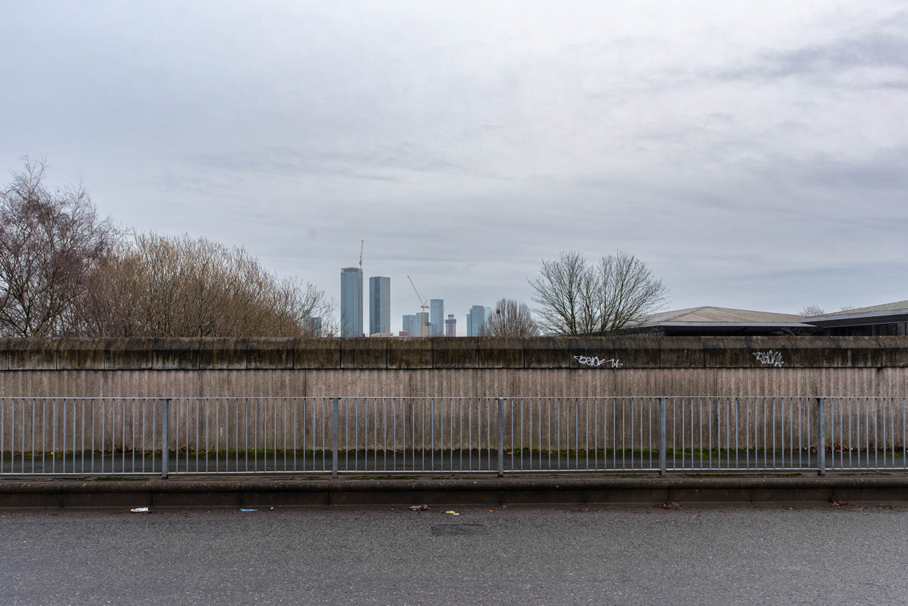 Straight-on view of a tall concrete wall behind a roadside barrier, with distant high-rise buildings visible above the top edge