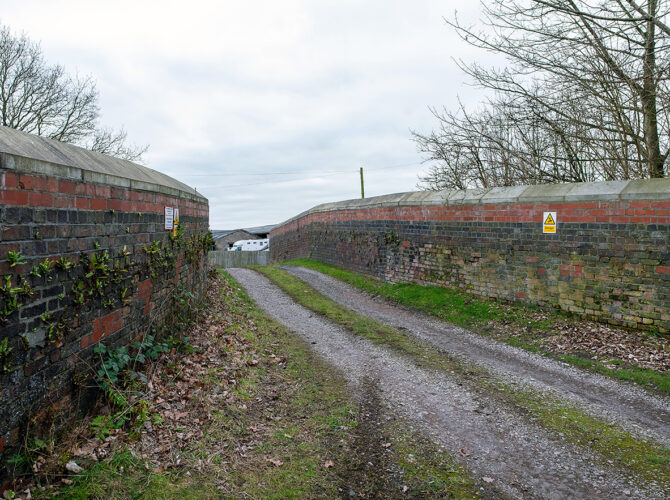A narrow gravel track runs over Newton’s Bridge, flanked by low brick parapet walls. Vegetation grows along the edges, and the enclosed passage emphasises its use as a private farm access route.