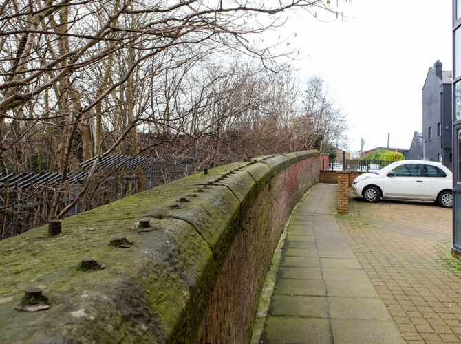Moss-covered railway wall curves past trees towards nearby flats and parked car