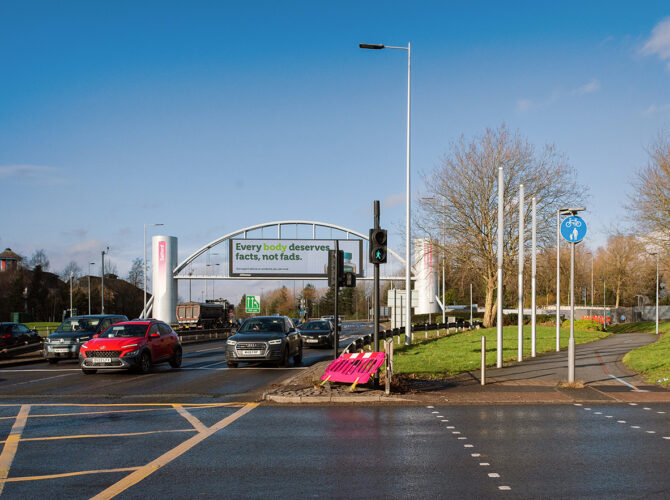 View across a multi-lane road with cars passing beneath a large white sign gantry, with pedestrian crossing lights and paths at the edge