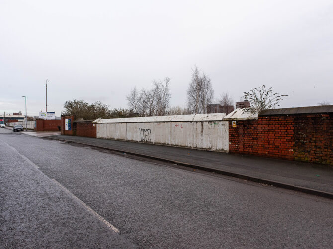 Broad view down a quiet road lined by a long retaining wall, with low industrial units, sparse traffic and overcast sky creating a subdued setting