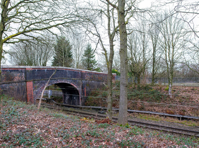A side view of Newton’s Bridge showing its rounded brick arch spanning an active railway line below. The bridge is set within a tree-lined cutting, with darkened brickwork from age and soot visible along the arch.