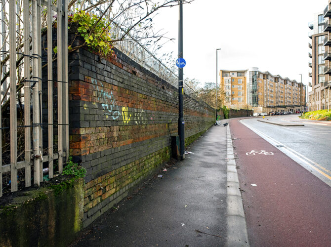 Long view of the old railway wall beside the road, with modern flats in the background.