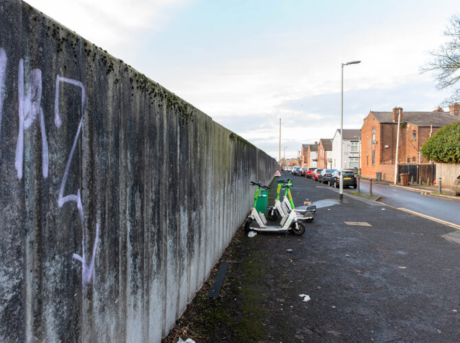 Close view along a tall concrete wall beside a pavement, with graffiti and parked e-scooters near residential houses