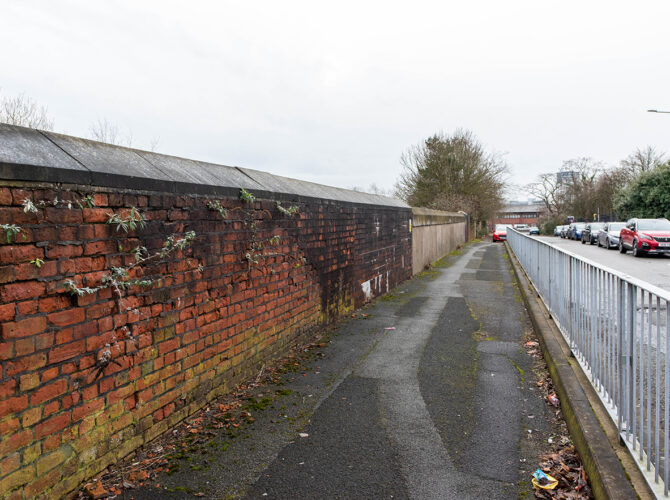 View along a narrow pavement with a red brick retaining wall on one side and a metal railing separating the road, with parked cars stretching into the distance