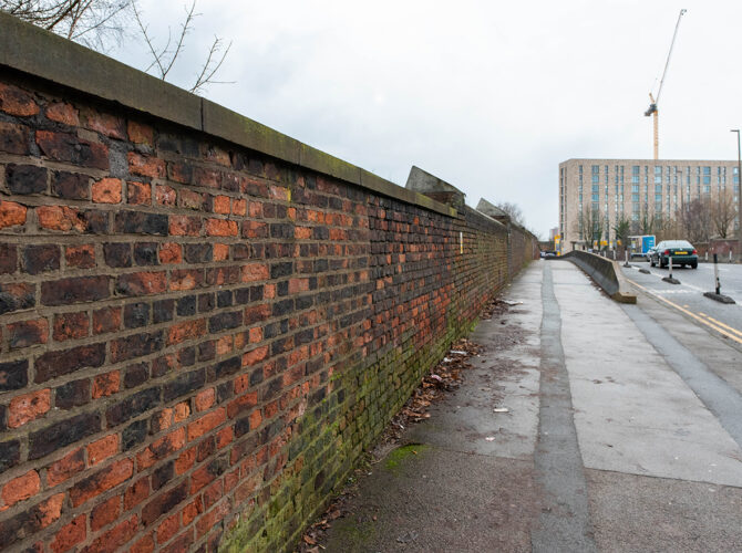 Perspective view along aged brick wall beside a quiet roadside pavement with passing traffic