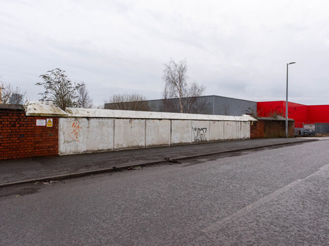 View along a wet roadside with a continuous retaining wall made of concrete panels and red brick sections, with industrial buildings and a large red warehouse in the background