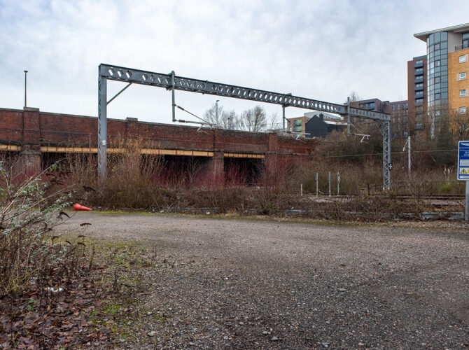 View of the line running under a low bridge with overhead gantry and scrubby vegetation