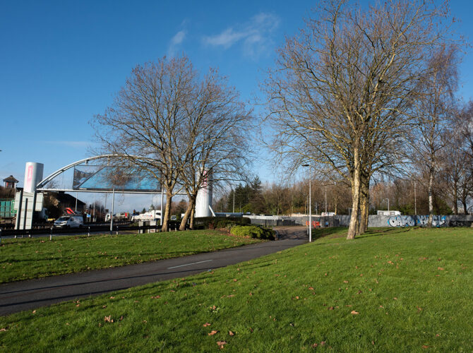 Curving pedestrian path across a grassy area beside the roundabout, with a large modern overhead sign structure spanning the road in the background