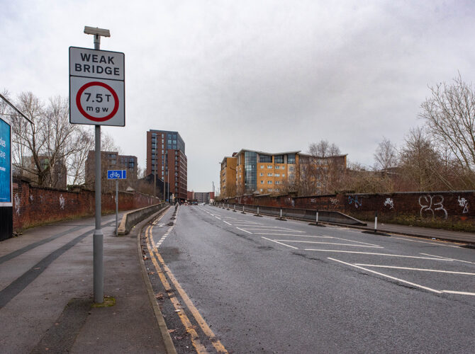 Street view with weight limit sign and continuous brick walls marking the raised railway route