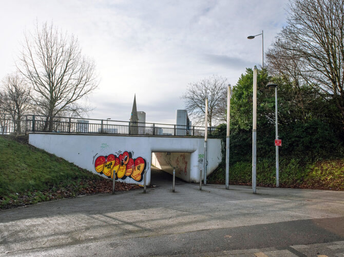 Entrance to a concrete pedestrian underpass set into a grassy embankment, with graffiti on the walls and railings and roadway above