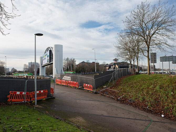 Pedestrian area beside the roundabout with barriers and fencing, with the bridge area screened from view by high panels