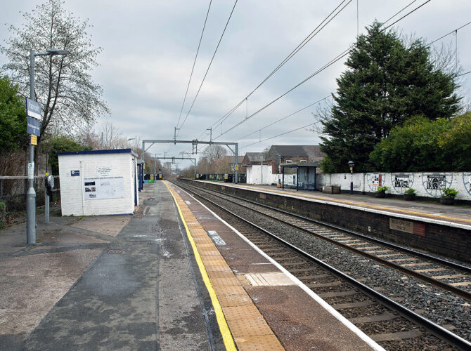 A view along the platform with electrified tracks, simple shelters and a white brick boundary wall.