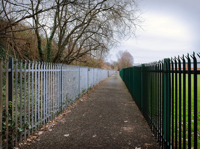A straight footpath between fences leads towards the hidden bridge beneath the railway embankment.
