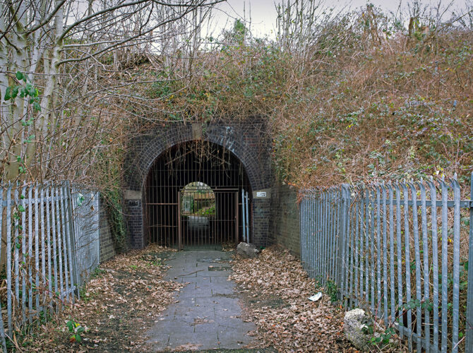 The bridge entrance framed by dark brickwork and overgrown vegetation, now secured with a metal gate.