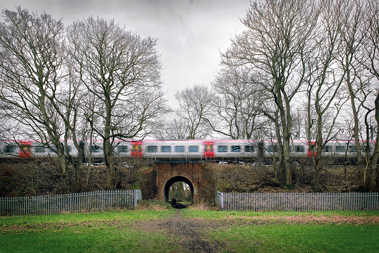 A red-and-white train crosses a low brick bridge above a narrow path running through an arched tunnel. Leafless trees and an overcast sky create a quiet, wintry atmosphere